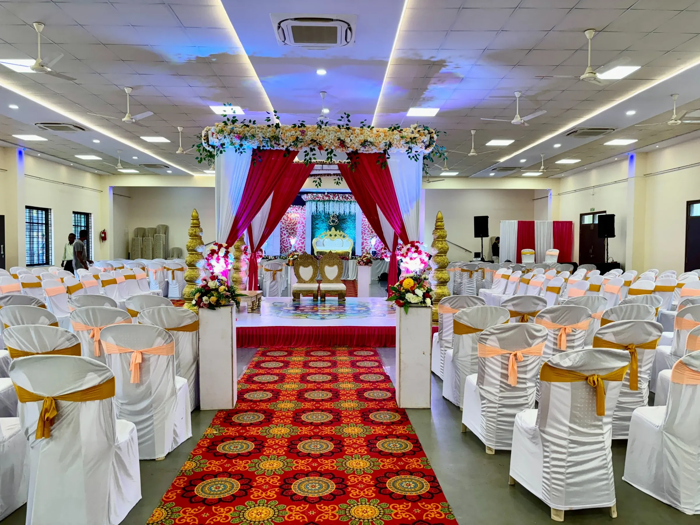 Wedding mandap with floral decor, white seating and a red patterned aisle in the hall at Yeshwant Hall, Ponda